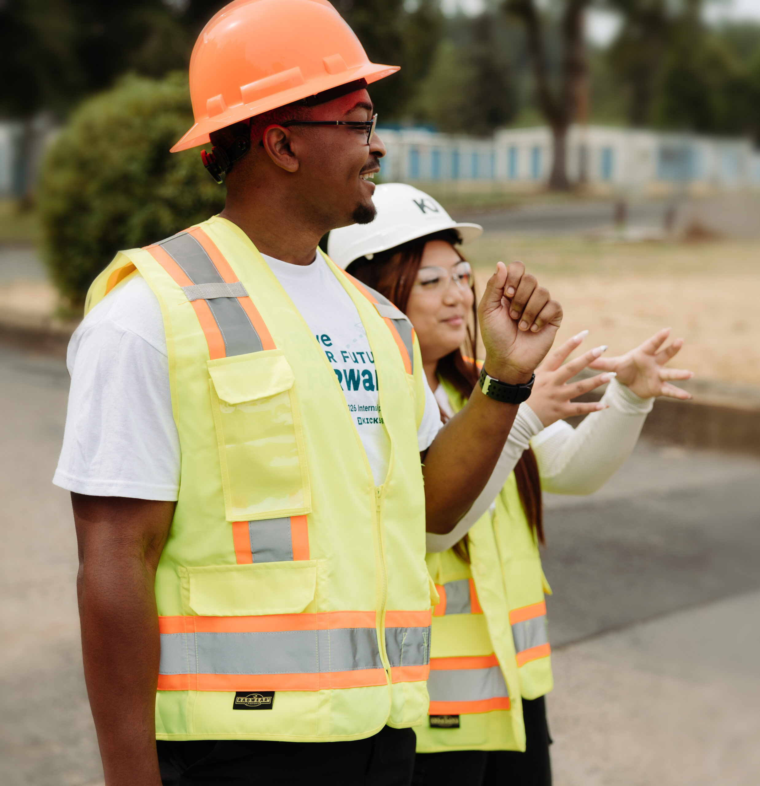 2 interns, on a guided tour of a job site, observe an area and appear to discuss it between one another. The interns gesture with their hands and smile as they converse. Both interns wear fluorescent vests and hardhats. The intern on the left has short brown hair, glasses, and is tall. The intern on the right has long brown hair, glasses, and is shorter.