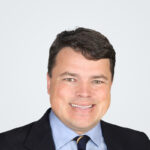 Headshot of Peter Symonds, a man with dark brown hair, in front of a light grey background.