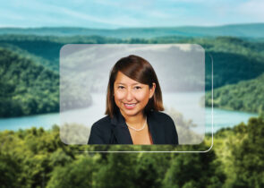 A headshot of Katty Fleming, a woman with brown hair, is centered on top of a photo of a landscape. The landscape setting is in a MidAtlantic state. It depicts rolling hills with green trees and a soft blue sky.