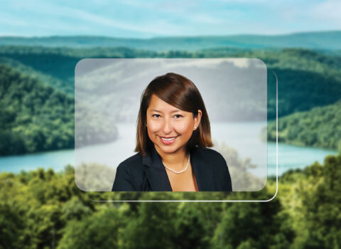 A headshot of Katty Fleming, a woman with brown hair, is centered on top of a photo of a landscape. The landscape setting is in a MidAtlantic state. It depicts rolling hills with green trees and a soft blue sky.