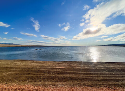 A landscape photo of part of the High Desert Water Bank in Southern California. A bank of water shimmers under a blue sky and is surrounded by earth-tone gravel.