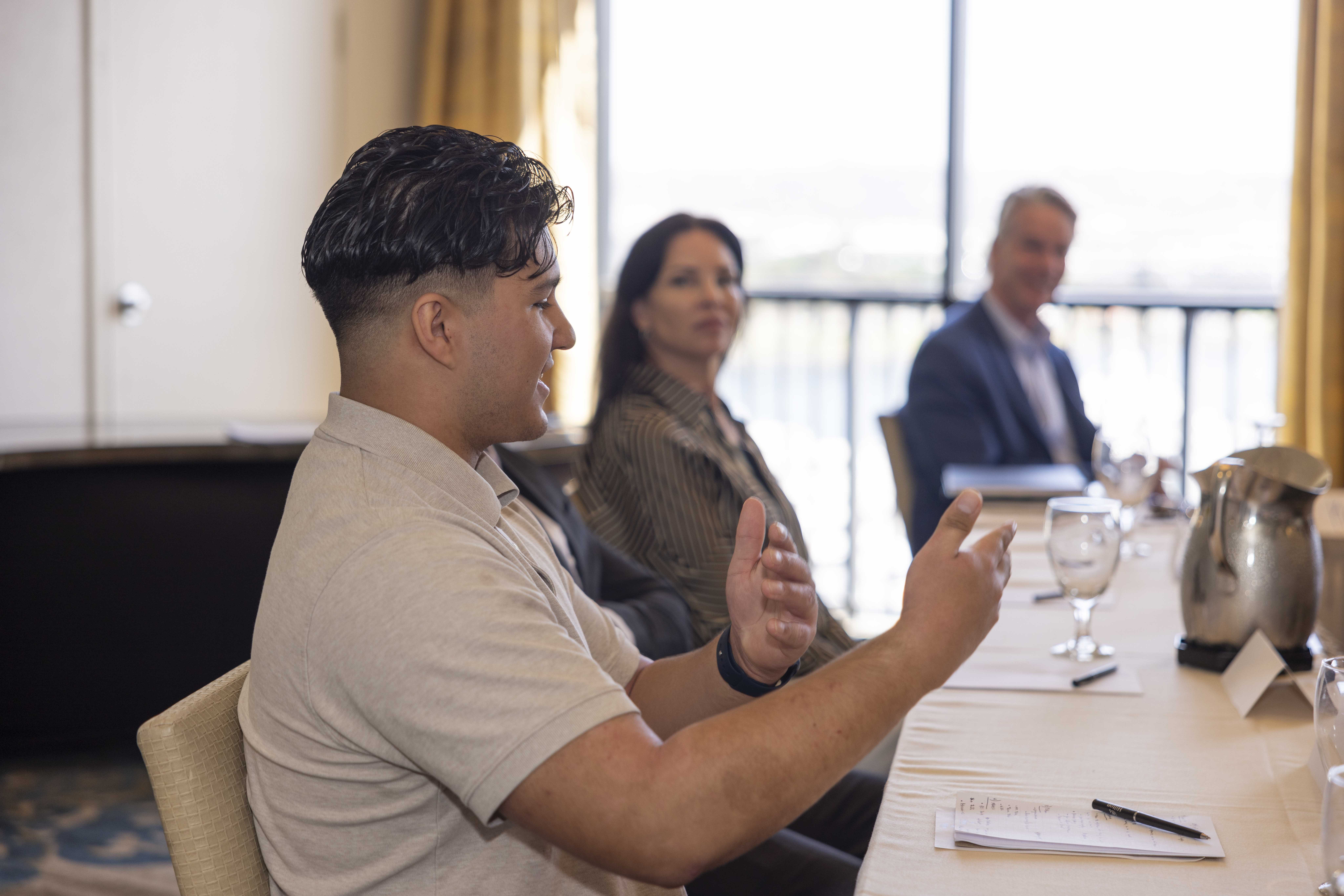 A KJ intern in a beige polo shirt gestures with their hands while explaining a thought to a group. The group sits in a conference room partially lit by natural light coming in through the windows.