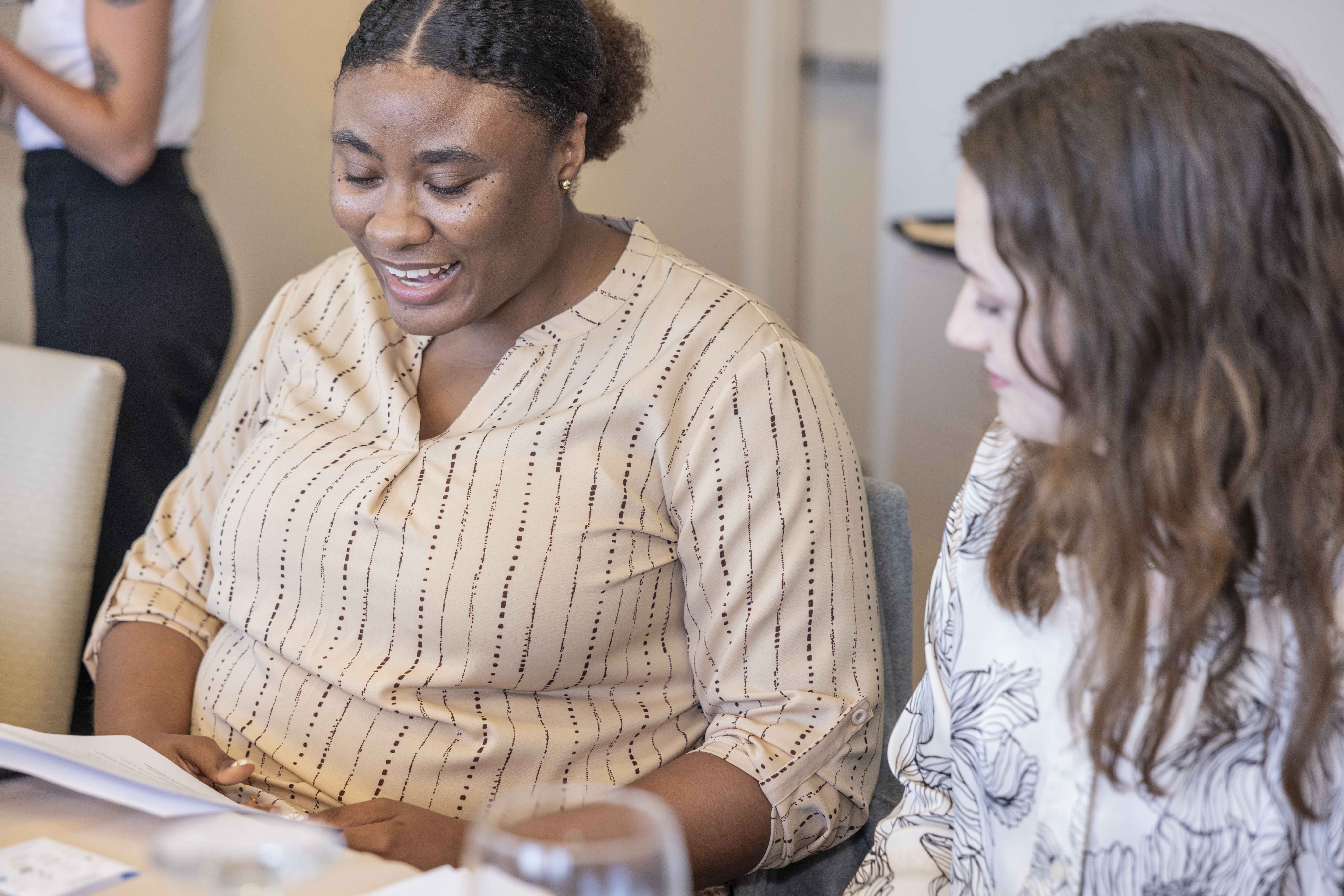 Two KJ interns, one in a cream blouse and the other in a white blouse, sit next to each other. The two interns examine a worksheet on a cloth table while smiling to each other.