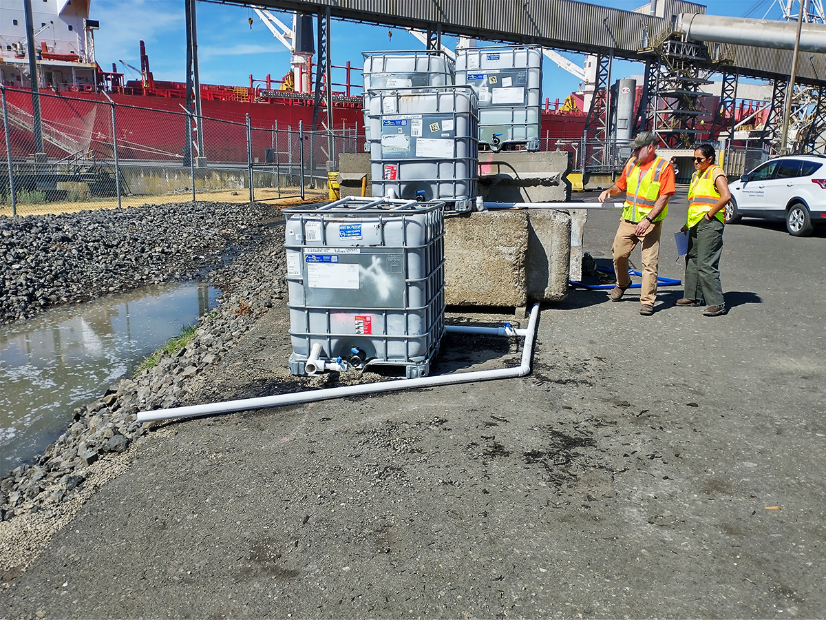 A KJ Intern and employee examine water-related construction on a sunny day. The intern and employee wear yellow and orange safety vests and stand on a grey gravel ground under a blue sky.