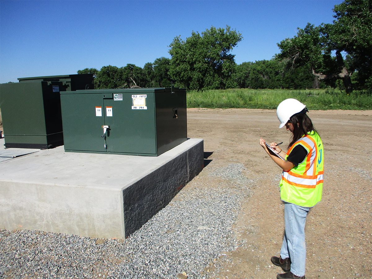 A KJ Intern examines construction on a sunny day. The intern wears a yellow and orange safety vest and stands on a grey gravel ground under a blue sky.