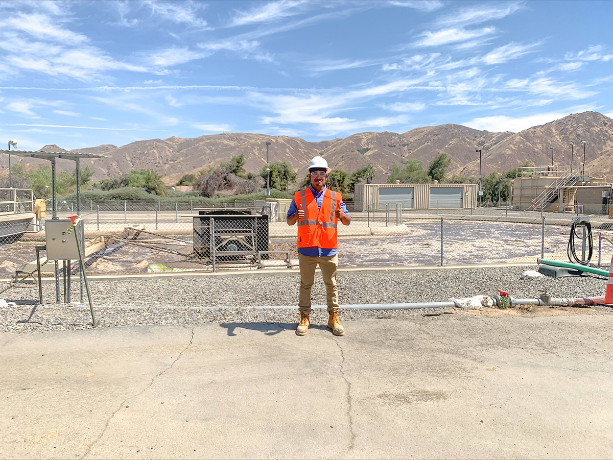 A KJ Intern stands in a water-related construction area holding two thumbs up. The intern wears an orange safety vest and stands on a tan cement ground under a blue sky on a sunny day.