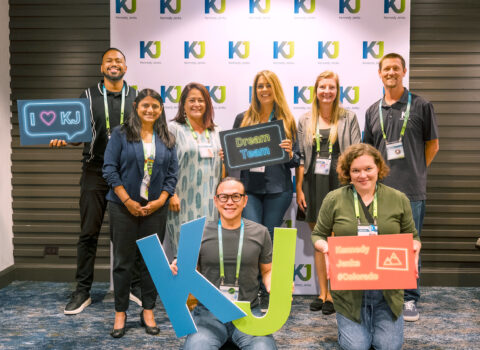 8 KJ employees pose for a picture at a company conference. The employees are men and women of various ages, wearing business casual attire. They stand in front of backdrop with a blue and green KJ logo pattern on it. This image relates to employee ownership.