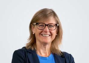 Katie Jones, Chief Strategy and Growtn Officer, Professional Headshot. A person with medium-length blond-brown hair, wearing glasses, a suit jacket, and a blue shirt.