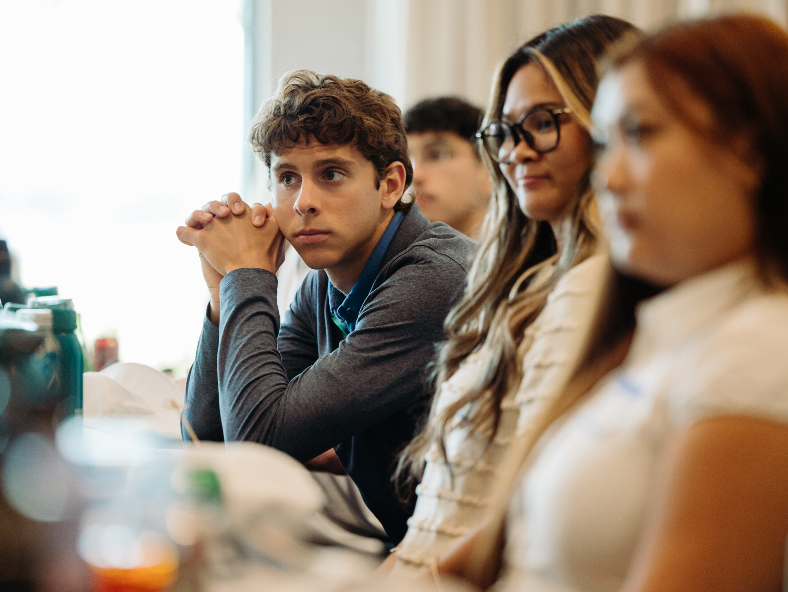 Interns sit at a conference table listening to a presentation. A person with short, brown curly hair and a grey sweater is in focus and appears to be listening intently to a presenter.
