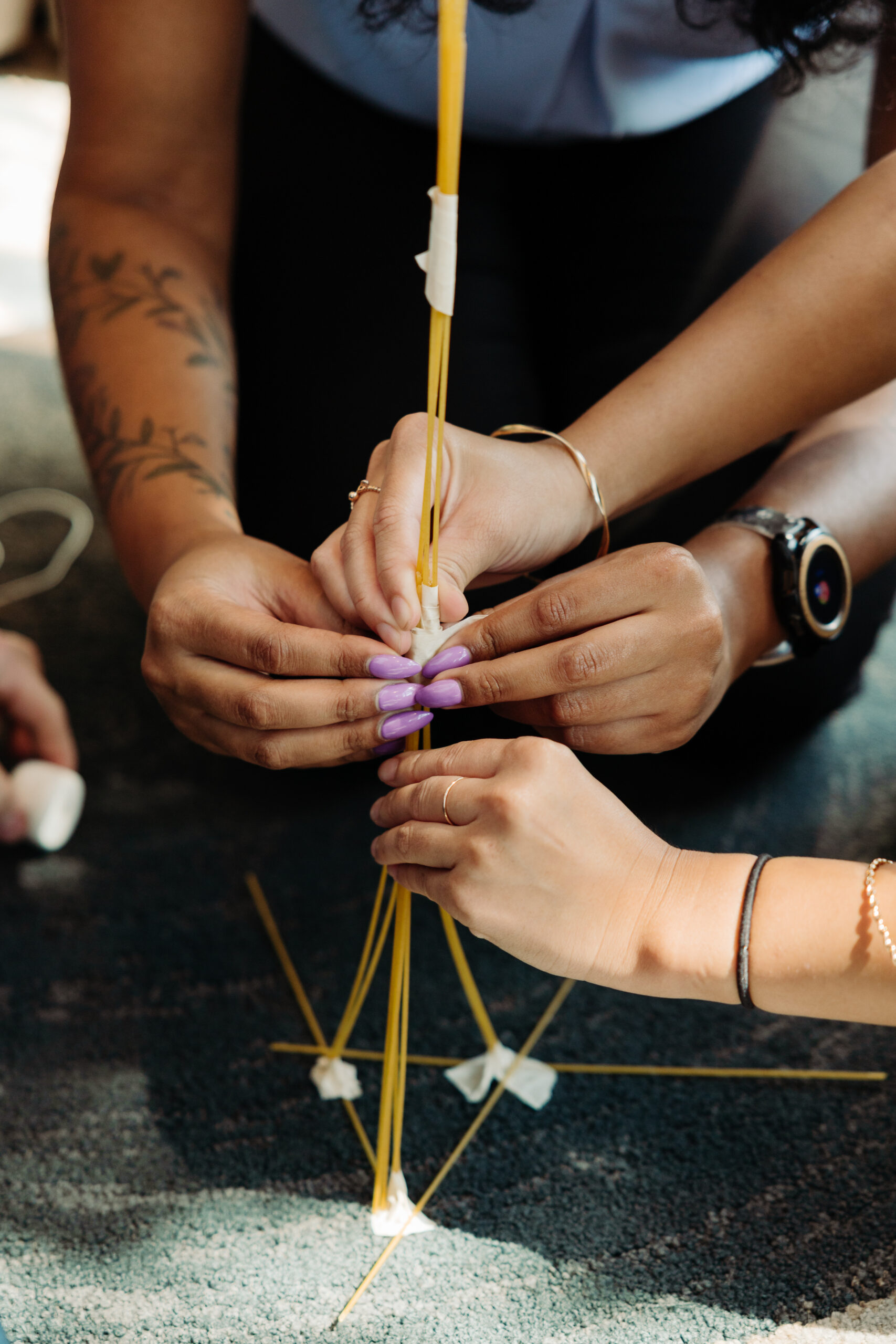 A close-up of interns' hands working to assemble a tall structure made out of pasta noodles and marshmallows - a team-building exercise.
