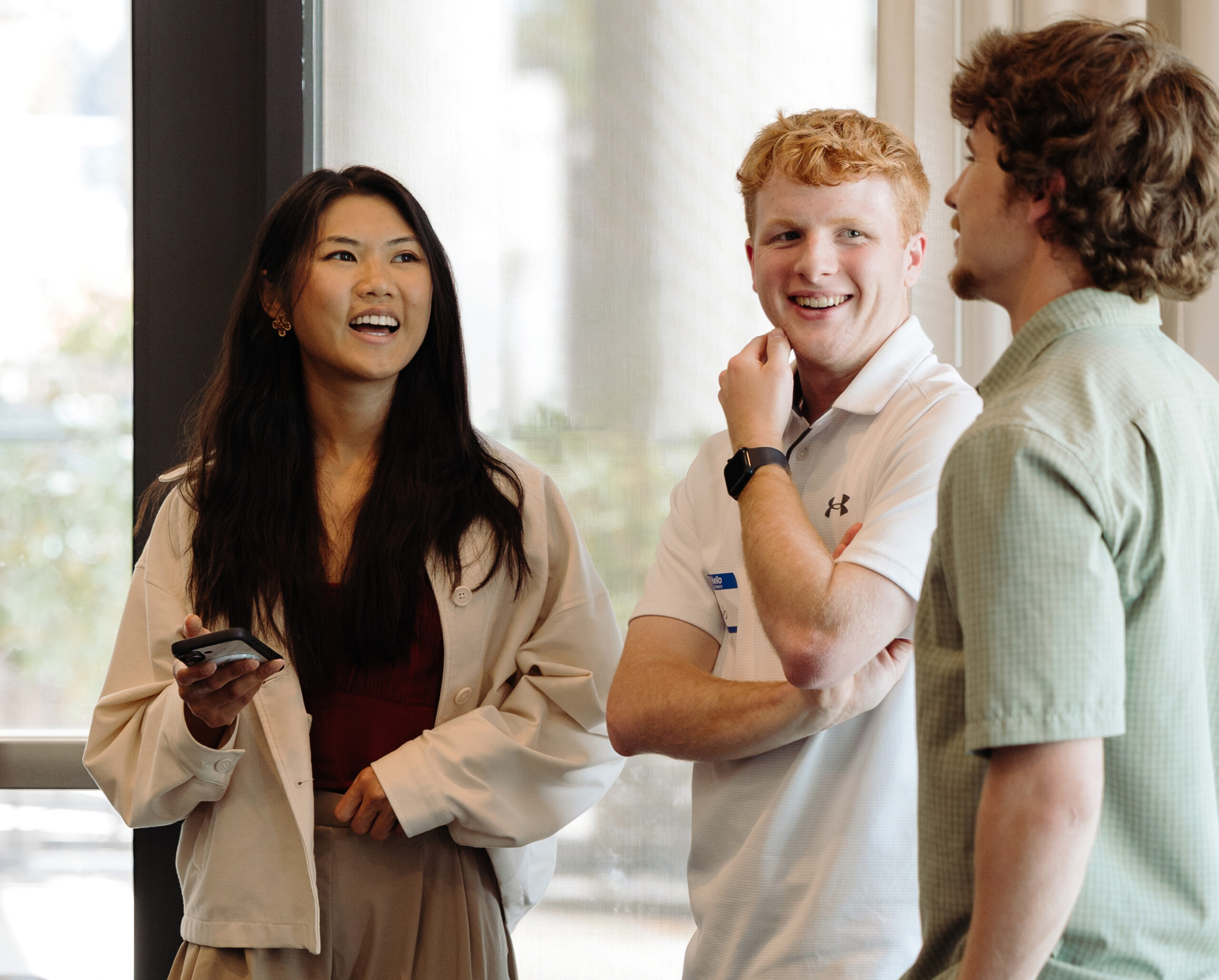 3 interns stand in a conference room talking, smiling, and laughing. The intern on the left has long dark brown hair and a cardigan on. The intern in the middle has short red hair and is wearing a white polo. The intern on the right has short brown hair, facial hair, and wears a light green polo.