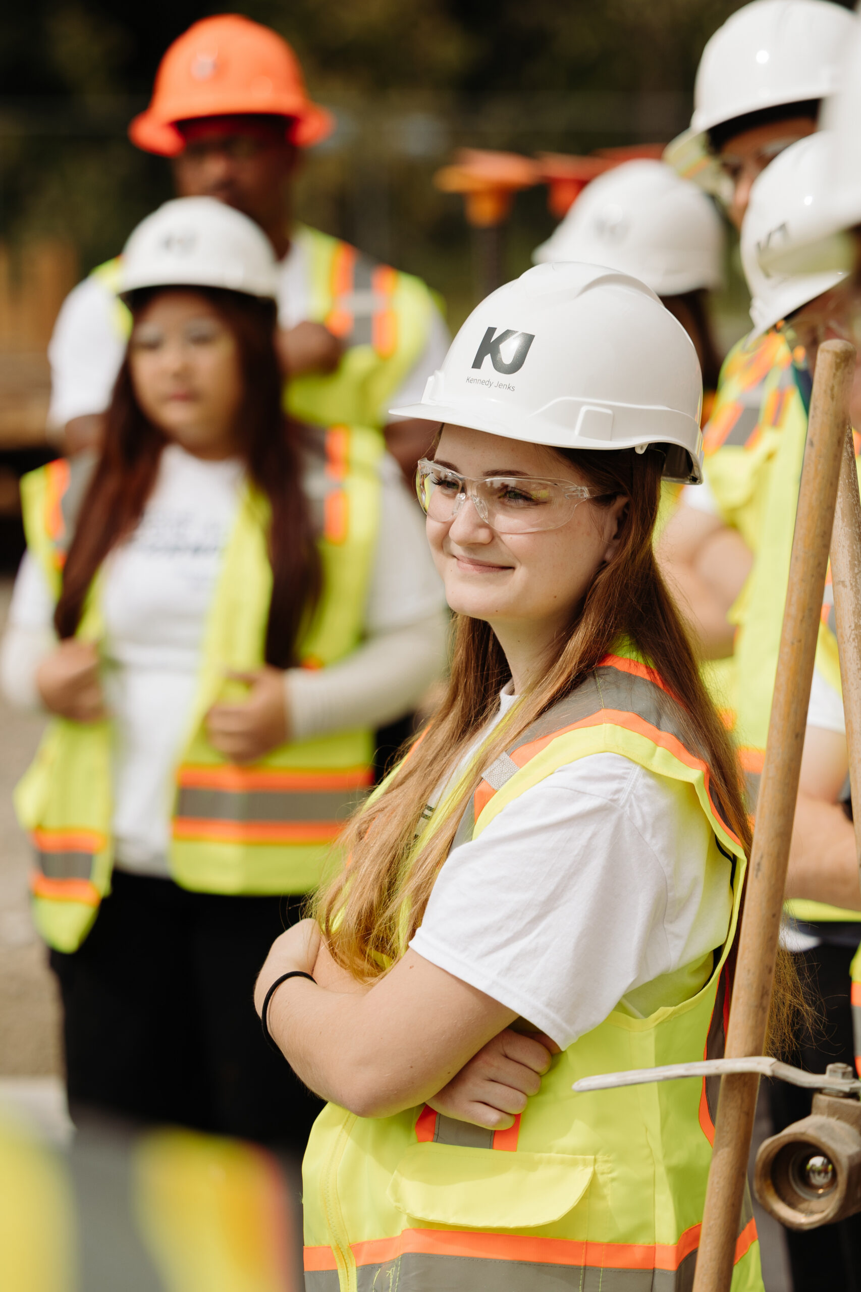Multiple interns, wearing fluorescent vests and hardhats, take a tour of a job site. 1 intern, with long brown hair and a white shirt, who is smiling, is in focus.