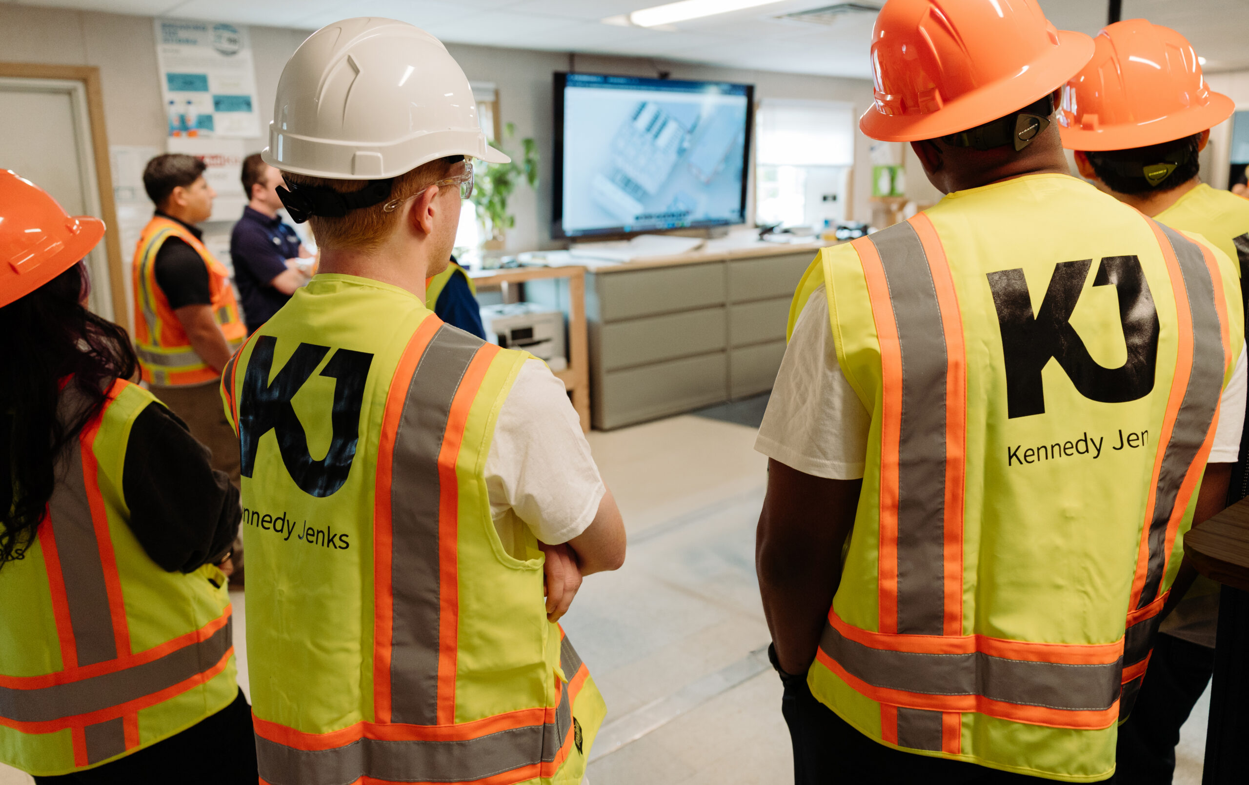 4 interns, wearing fluorescent vests and hardhats, stand in a room looking at a monitor displaying job site plans.