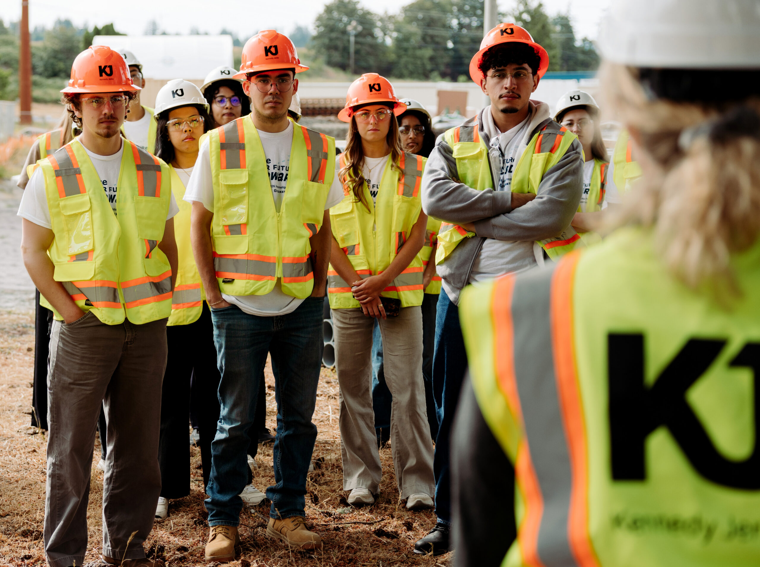 A group of interns, wearing fluorescent safety vests and hardhats, take a tour of a construction site. They look on as a KJ employee explains KJ's work at the site.