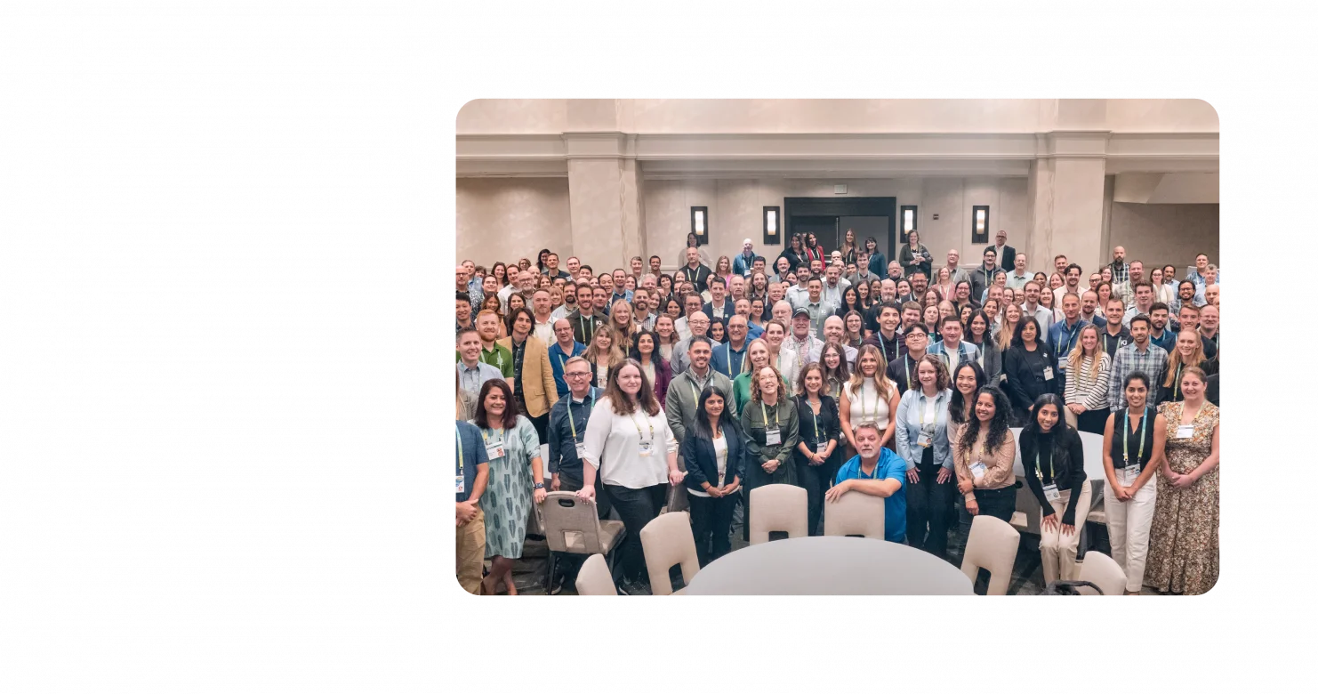 A large group of KJ employees pose for a photo at TechEx 2024. The employees are dressed in business casual clothing and stand in a cream-colored banquet room.