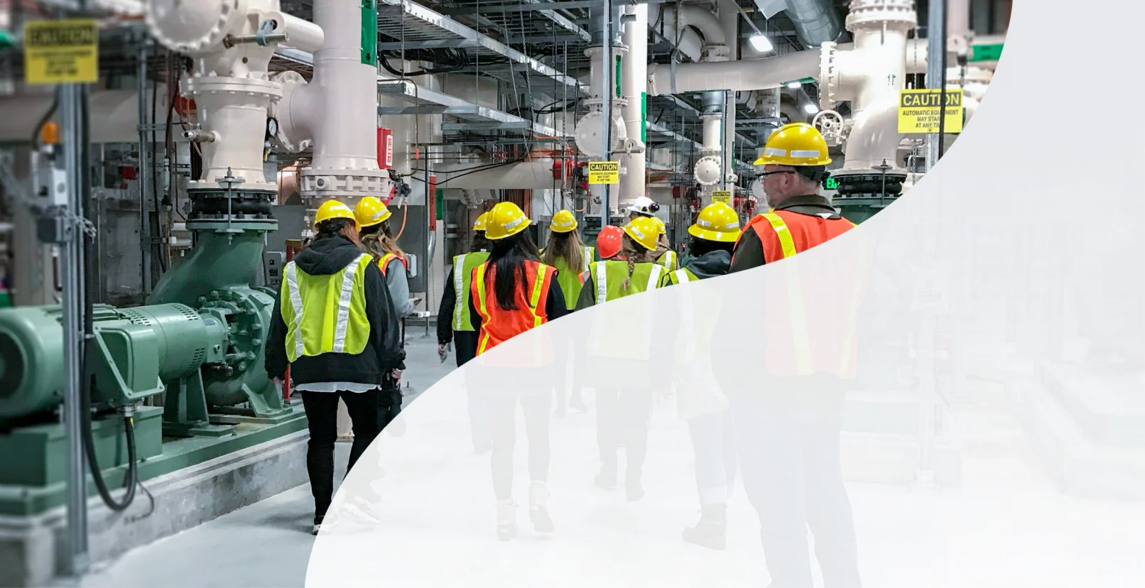 KJ employees explore a water treatment plant. The employees wear yellow hardhats, yellow and orange safety vests, are walking away from the camera, and surrounded by white and grey pipe structures.