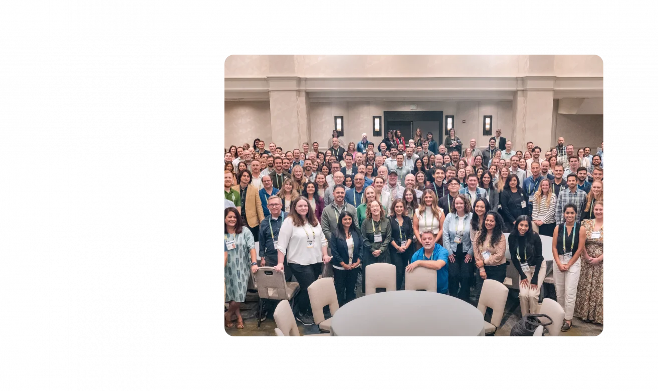 A large group of KJ employees pose for a photo at TechEx 2024. The employees are dressed in business casual clothing and stand in a cream-colored banquet room.