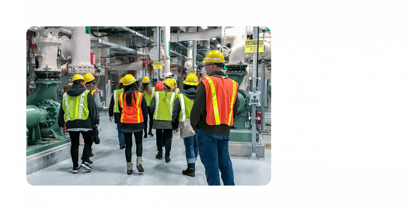 A handful of KJ employees walk through a water facility. They are wearing dark clothes with neon hard hats and neon vests on top.