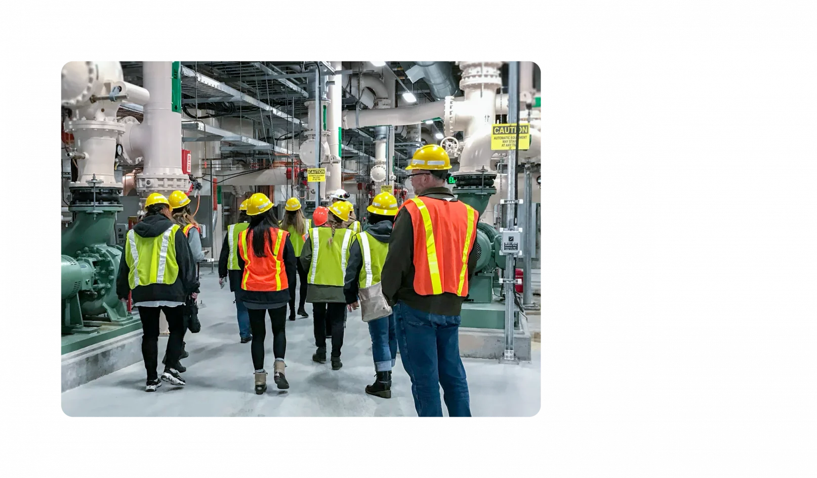 A handful of KJ employees walk through a water facility. They are wearing dark clothes with neon hard hats and neon vests on top.