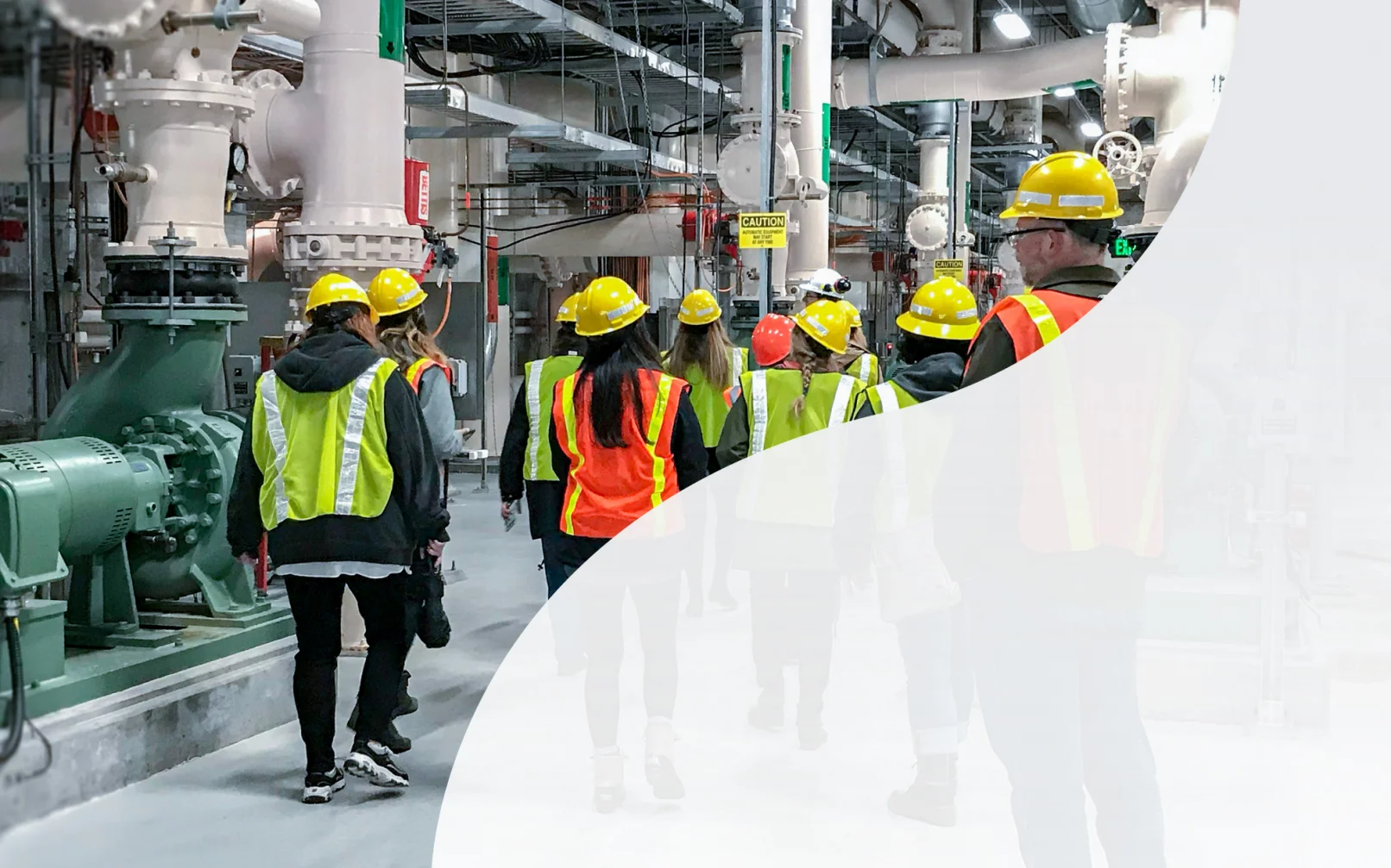 KJ employees explore a water treatment plant. The employees wear yellow hardhats, yellow and orange safety vests, are walking away from the camera, and surrounded by white and grey pipe structures.