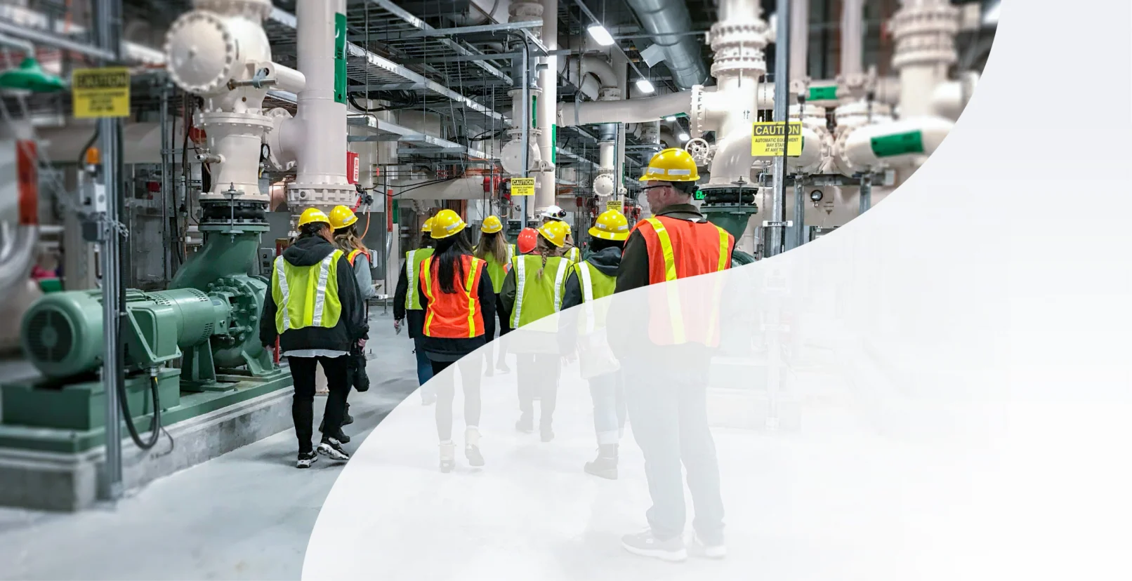 KJ employees explore a water treatment plant. The employees wear yellow hardhats, yellow and orange safety vests, are walking away from the camera, and surrounded by white and grey pipe structures.