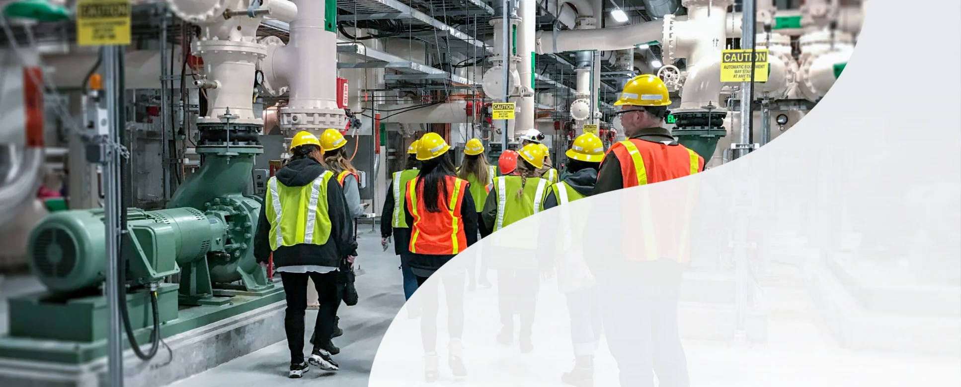 KJ employees explore a water treatment plant. The employees wear yellow hardhats, yellow and orange safety vests, are walking away from the camera, and surrounded by white and grey pipe structures.