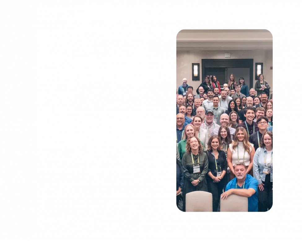 A large group of KJ employees pose for a photo at TechEx 2024. The employees are dressed in business casual clothing and stand in a cream-colored banquet room.